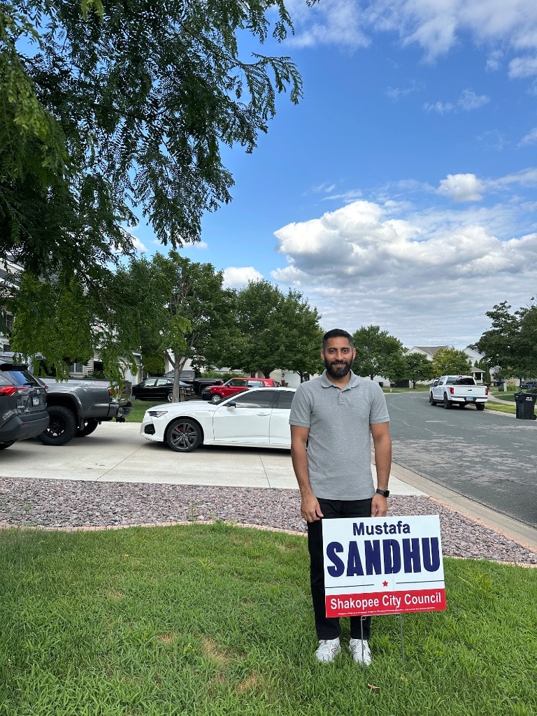 Mustafa Sandhu holding a campaign yard sign outdoors