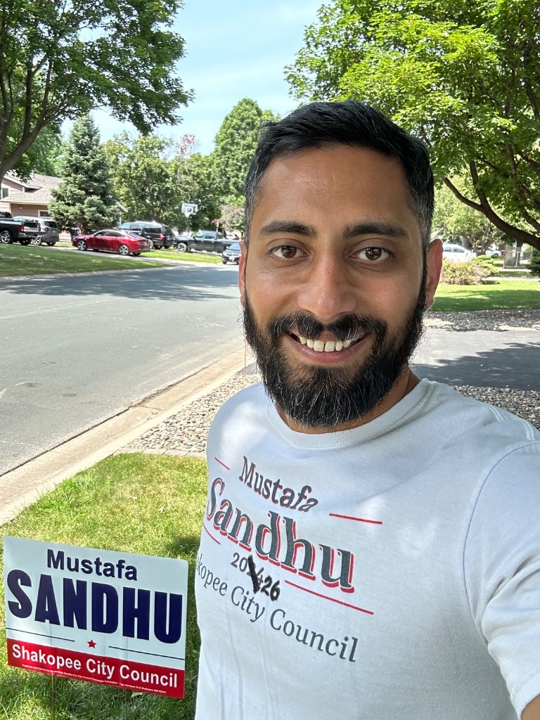 Mustafa Sandhu smiling with a political campaign sign outdoors